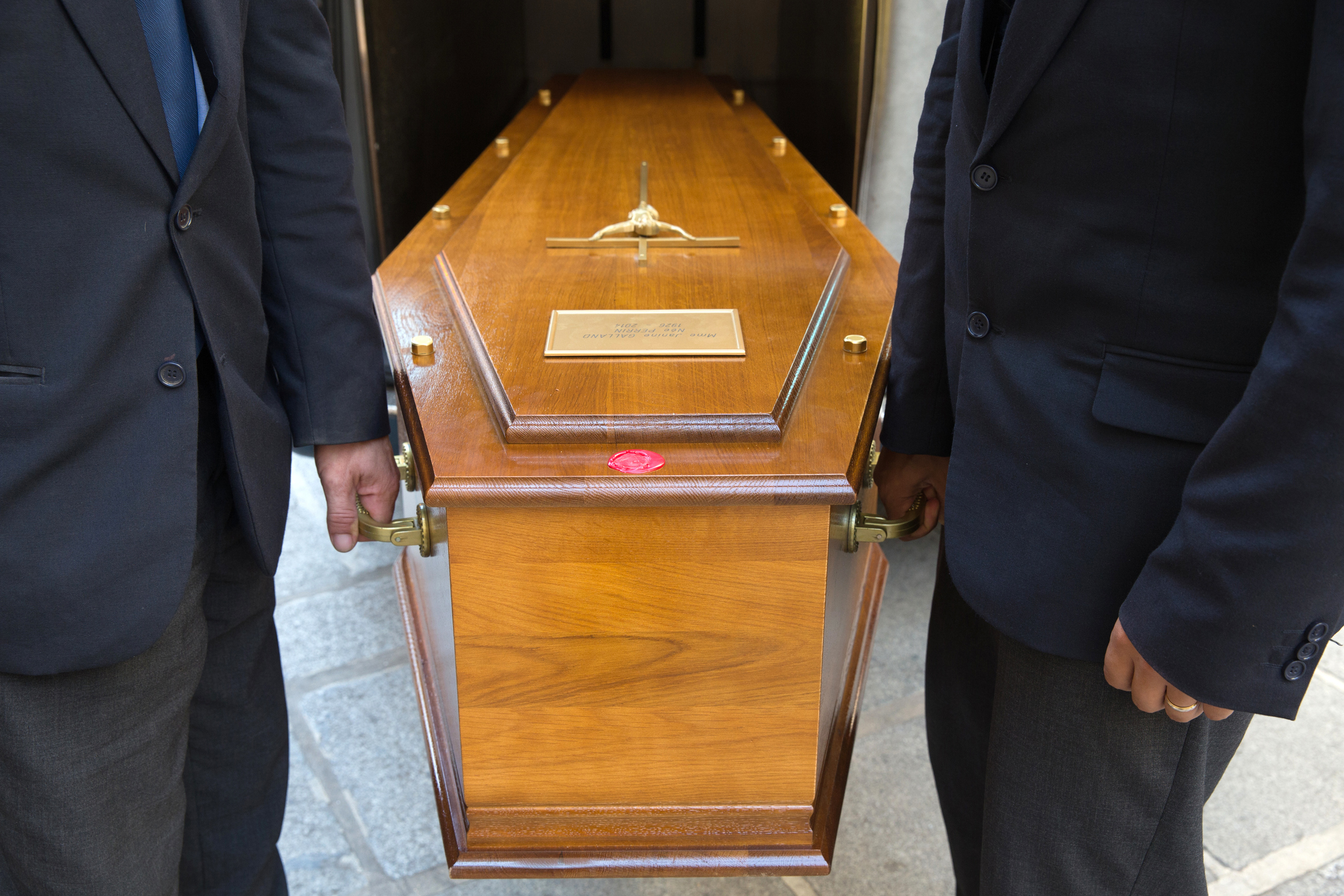 Two pallbearers in dark suits carry a coffin in a close-up photo