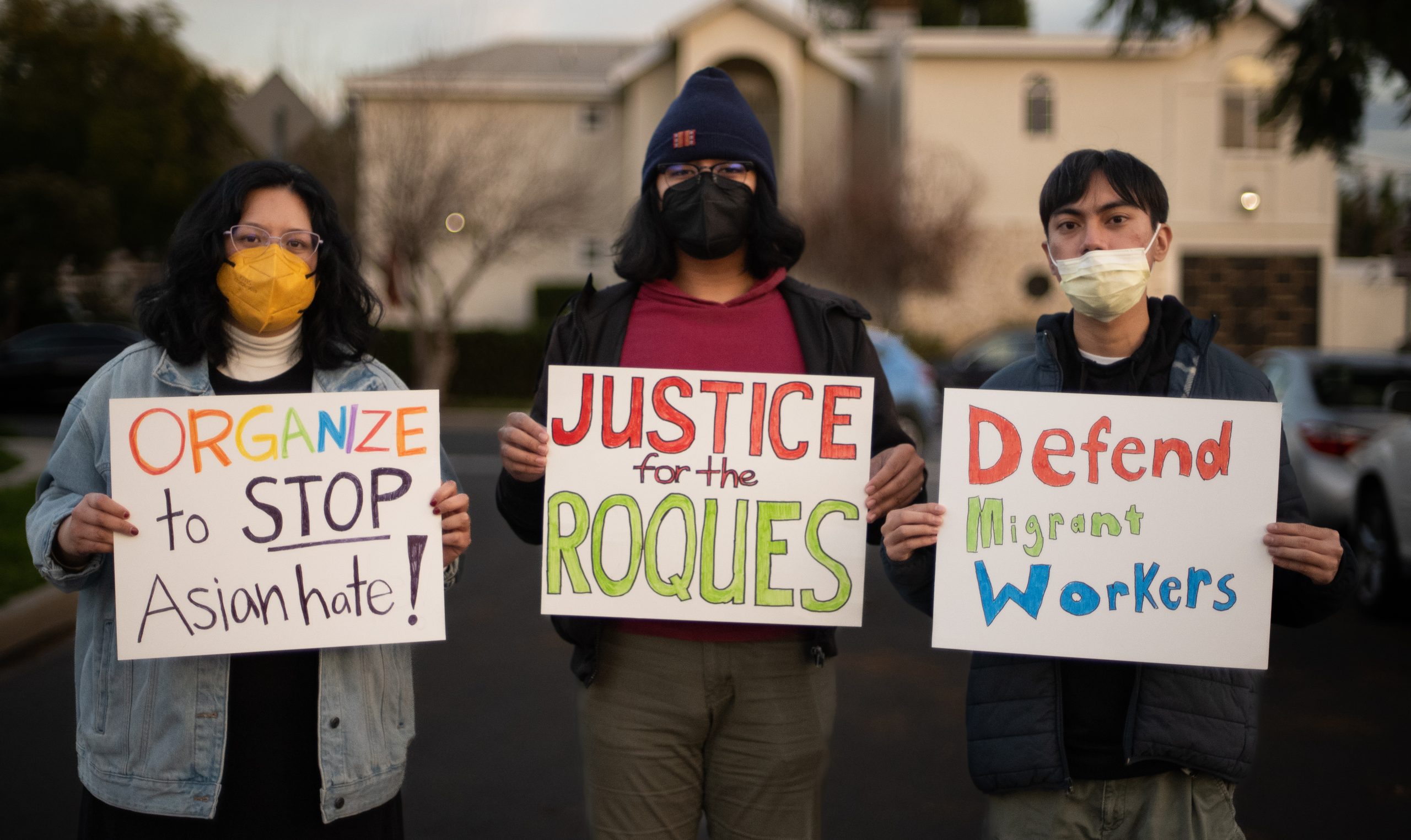 Three people stand outdoors, wearing face masks to protect against COVID, and holding signs. The signs say: "Organize to STOP Asian hate!" and "Justice for the Roques" and "Defend Migrant Workers"