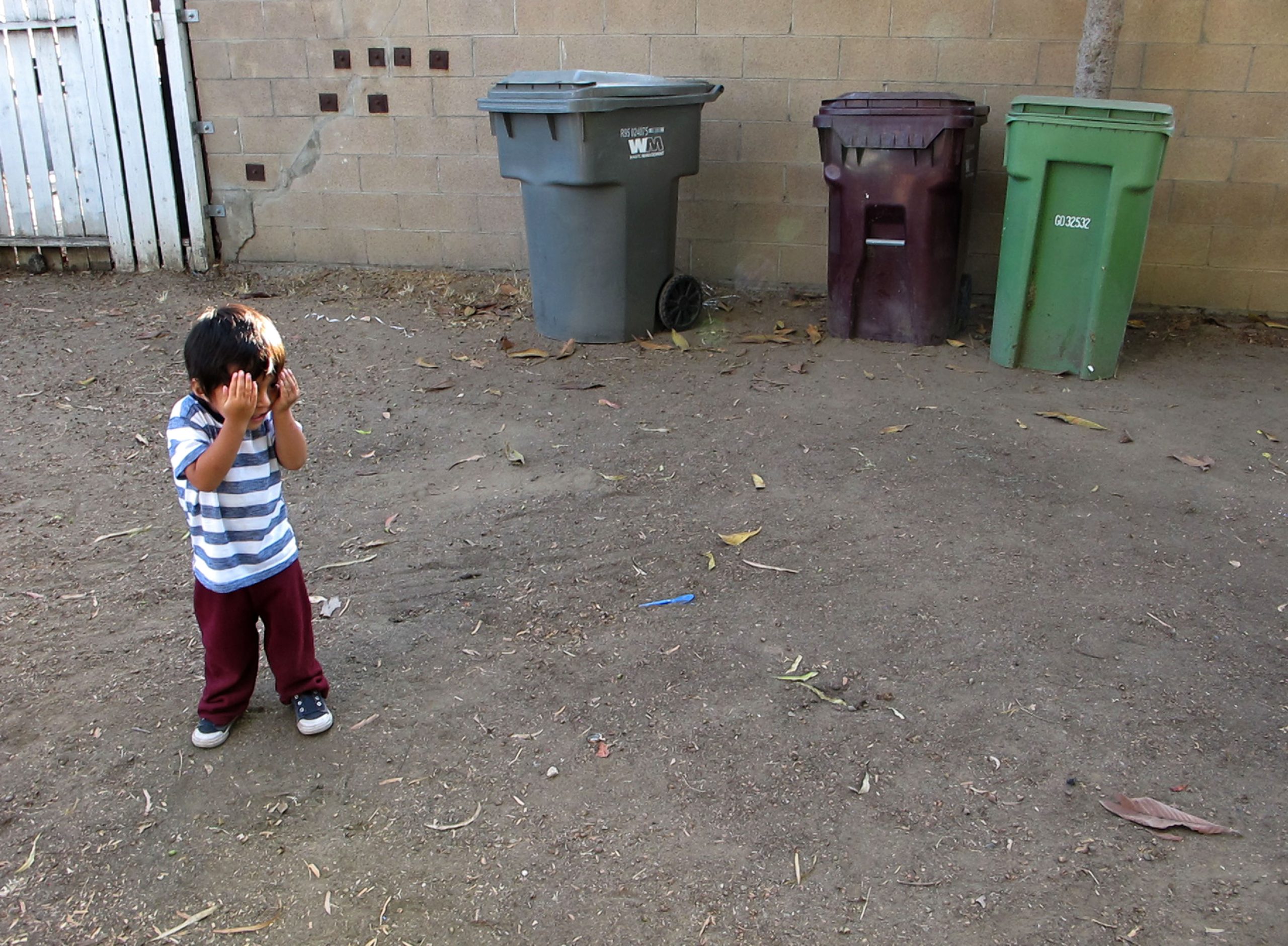 Ruben, 2, plays hide and seek in his front yard. He is surrounded by dirt on the ground and trash bins.