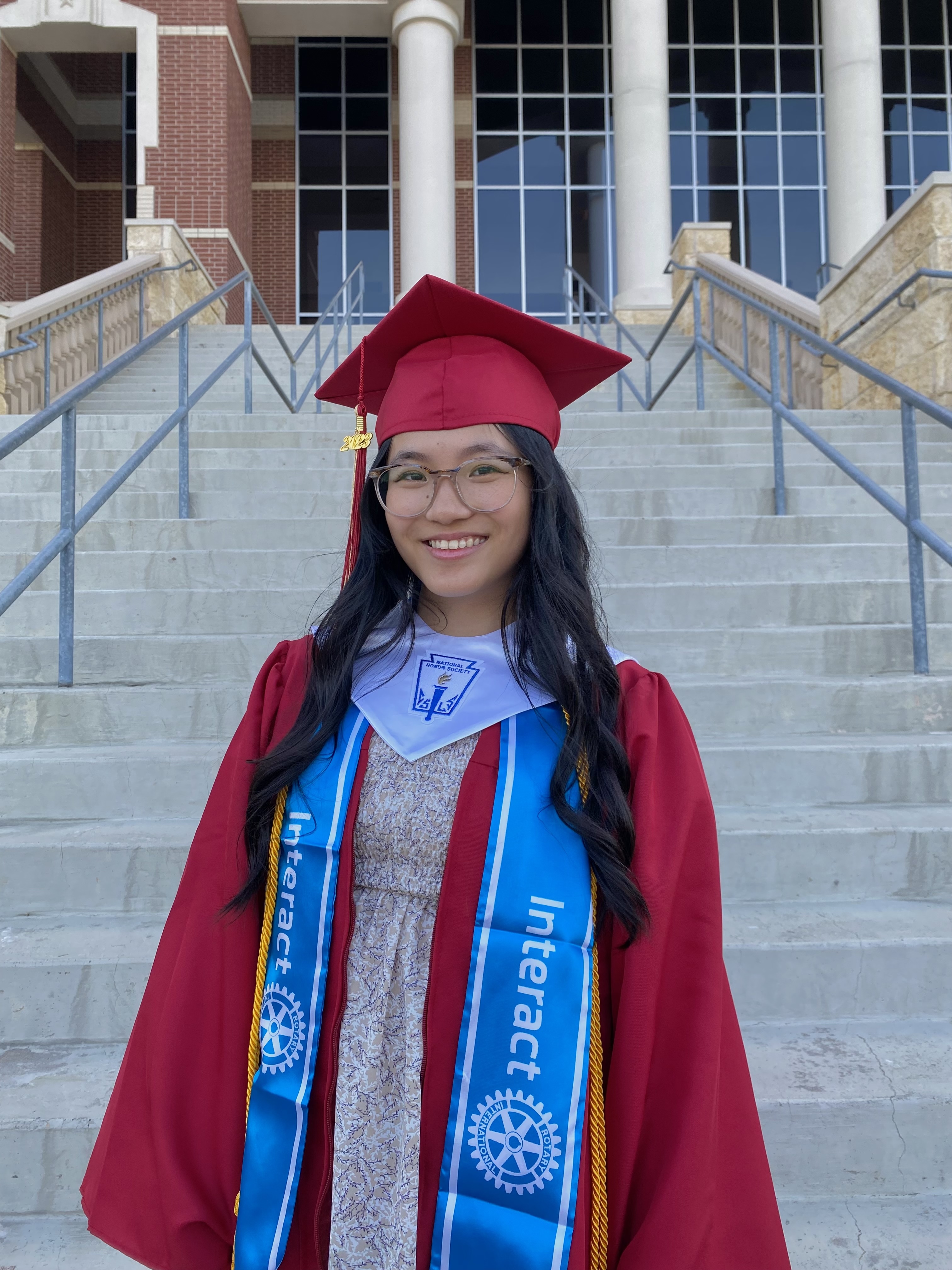 Victoria Zhang, in her graduation robe and cap, smiles on the exterior steps of the school