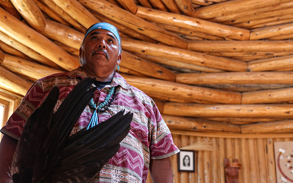 A Native American man with a blue headband, holds feathers for part of a traditional healing ceremony. He stands in a wooden hogan.