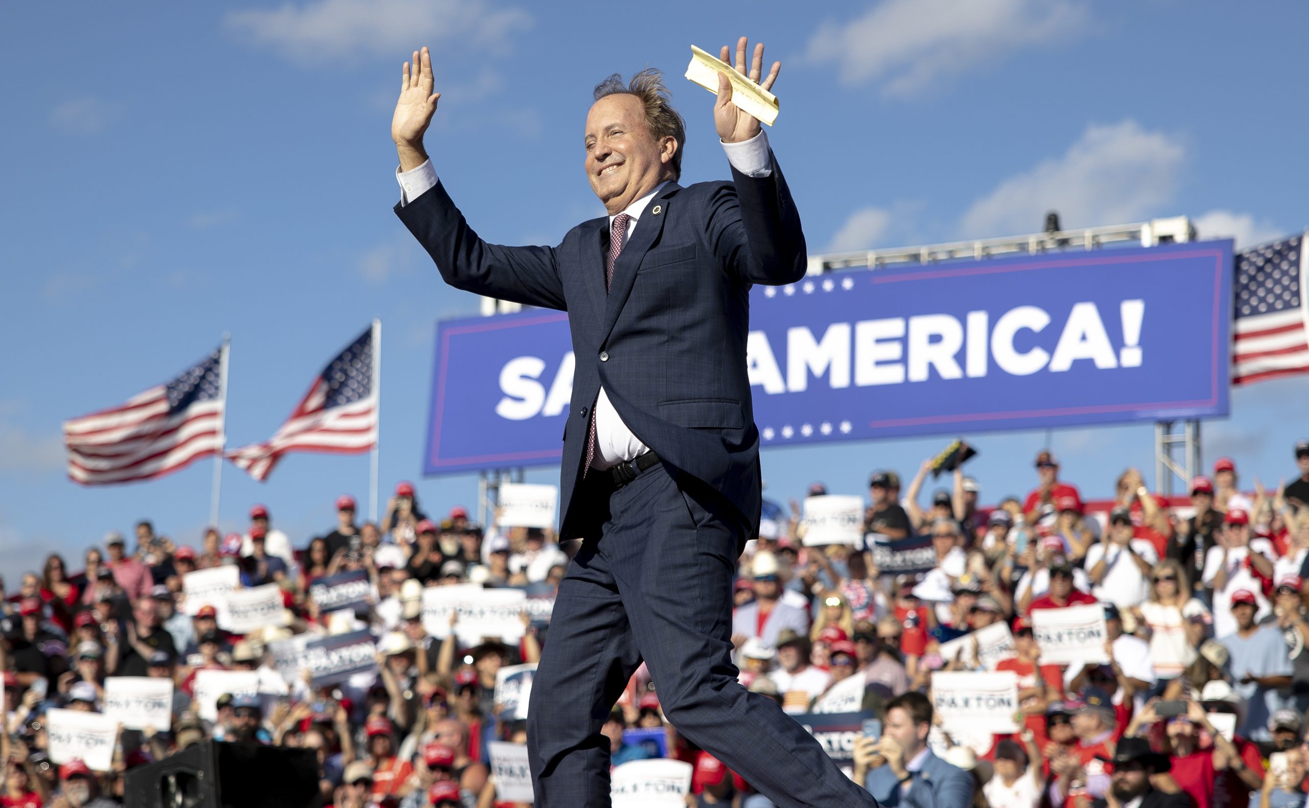 Ken Paxton waves to a crowd. A large banner behind him reads, “Save America!”