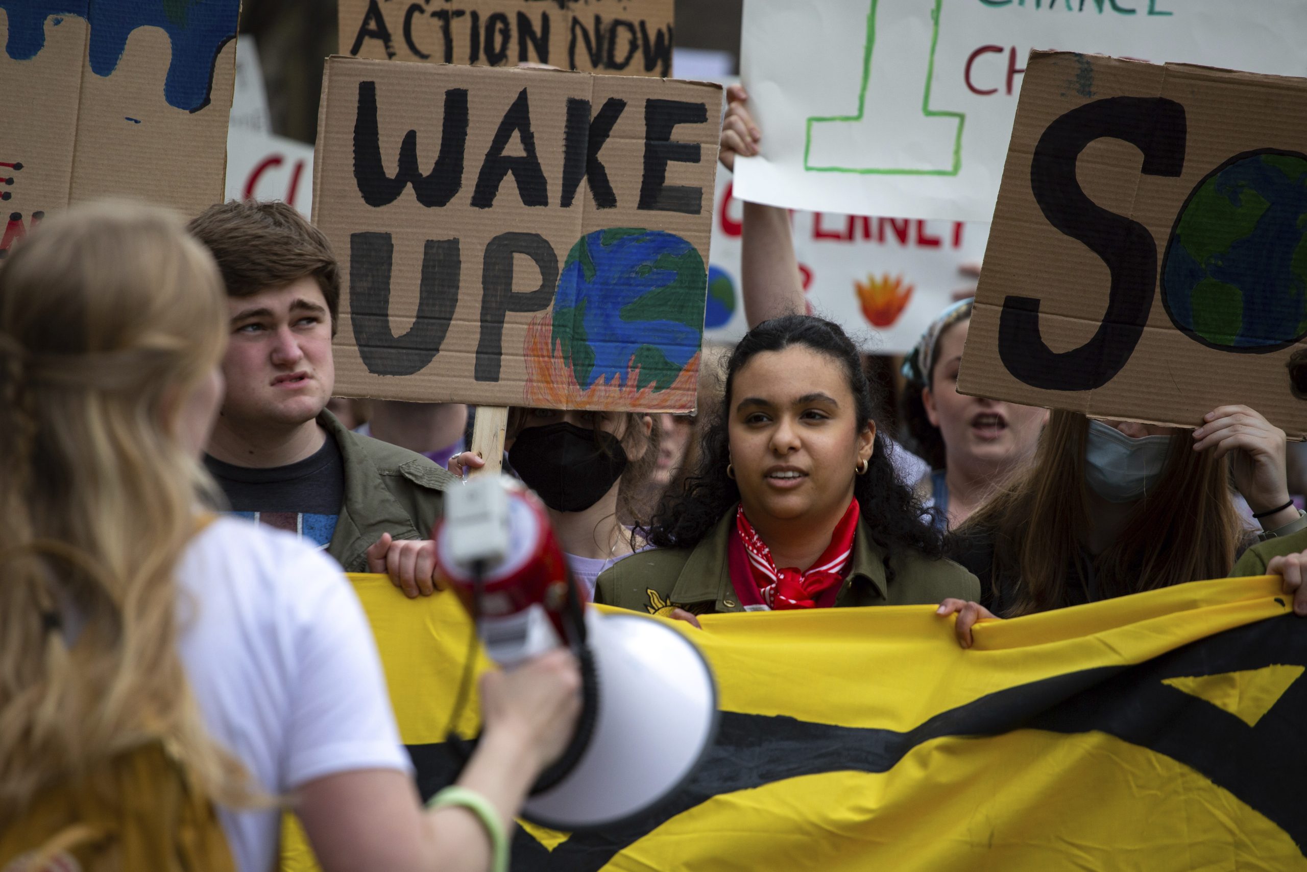 A group of people hold signs and banners. One sign reads, “Wake up,” and includes a drawing of the Earth in flames.