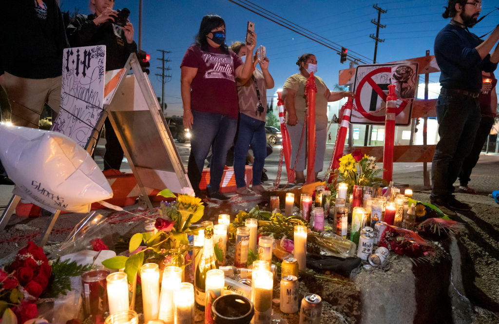 Mourners stand near a streetside memorial with candles and flowers.