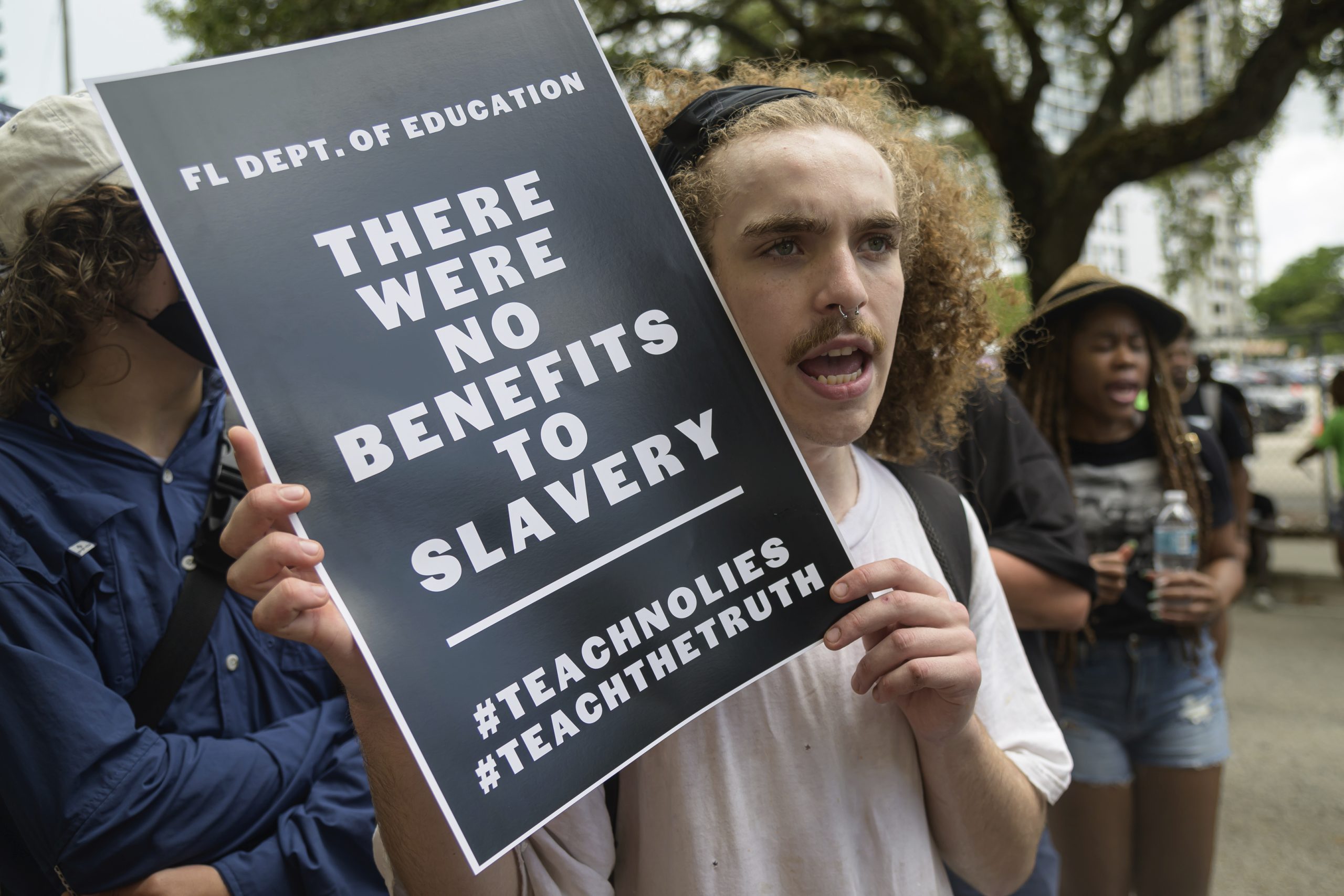 A young man holds a sign that reads: "FL Dept. of Education: There were no benefits to slavery. #teachnolies #teachthetruth"