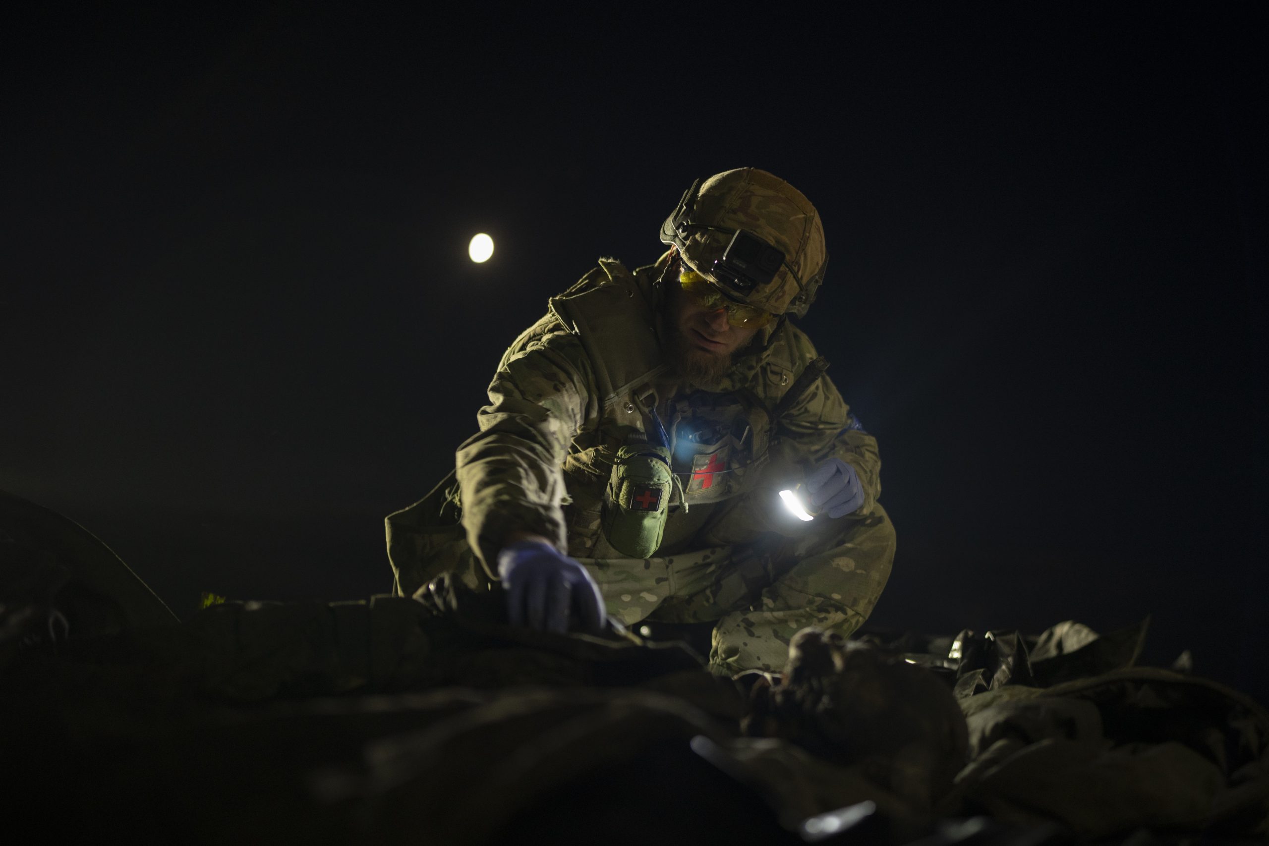 Oleksii Yukov, dressed in a camouflage uniform, holds a small light while examining a body. The body is shown in silhouette only.