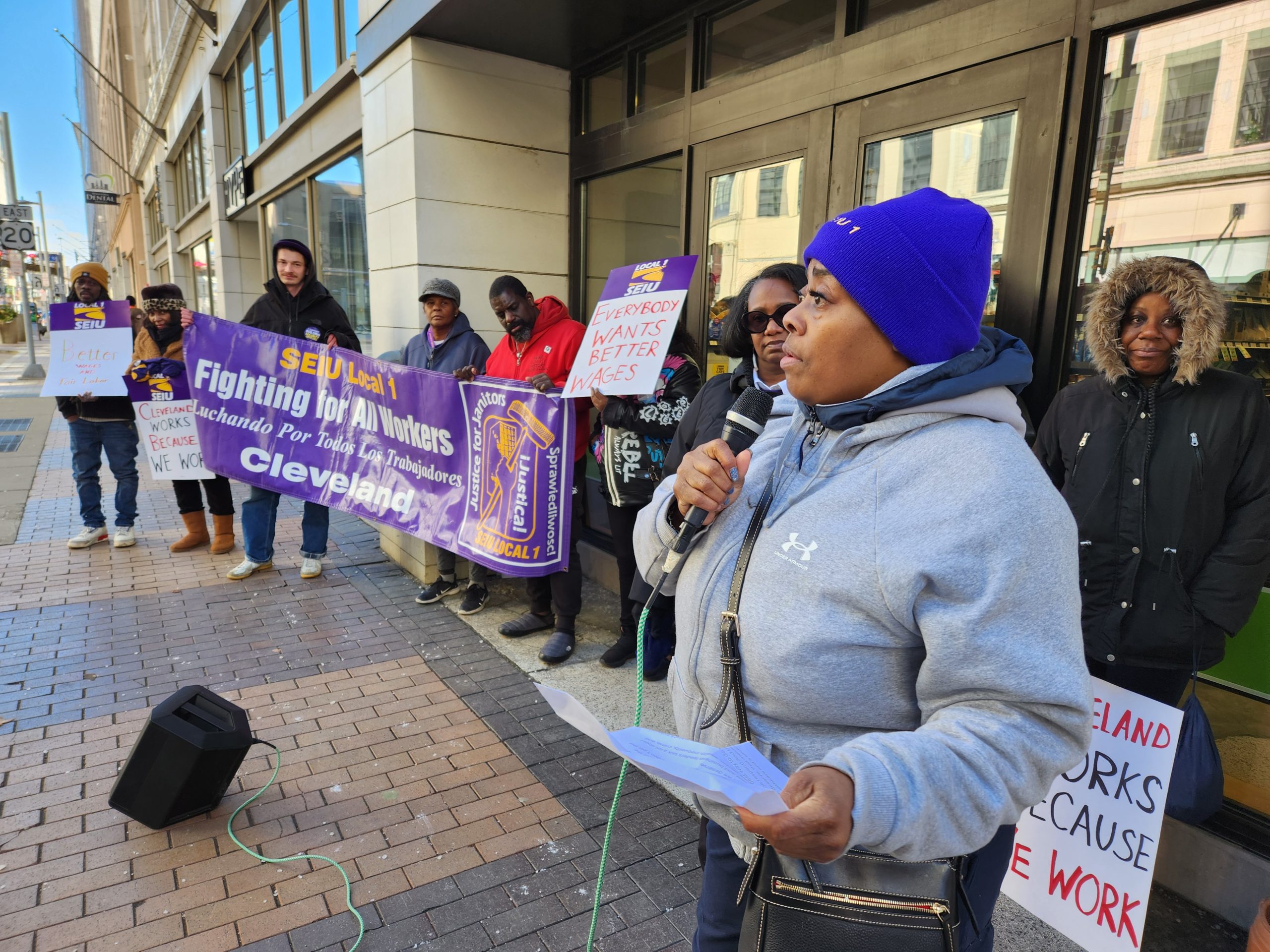 Janitors rally for higher wages in front of the offices of Downtown Cleveland, Inc. One woman speaks on a microphone. Everyone is dressed in coats. Some wear hats and gloves.