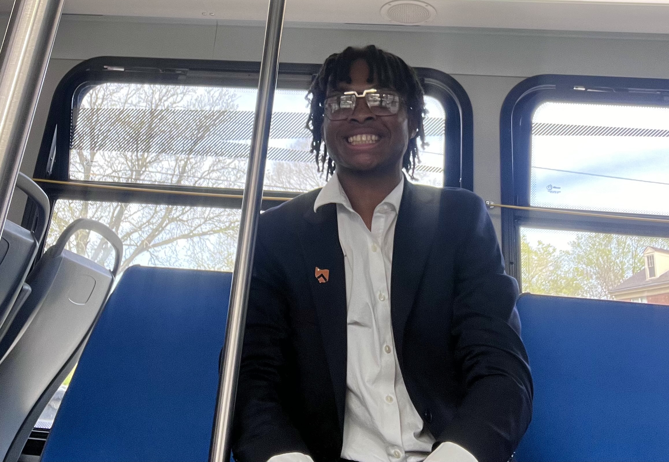 A young man sits on a blue bus seat in a suit jacket and is smiling.