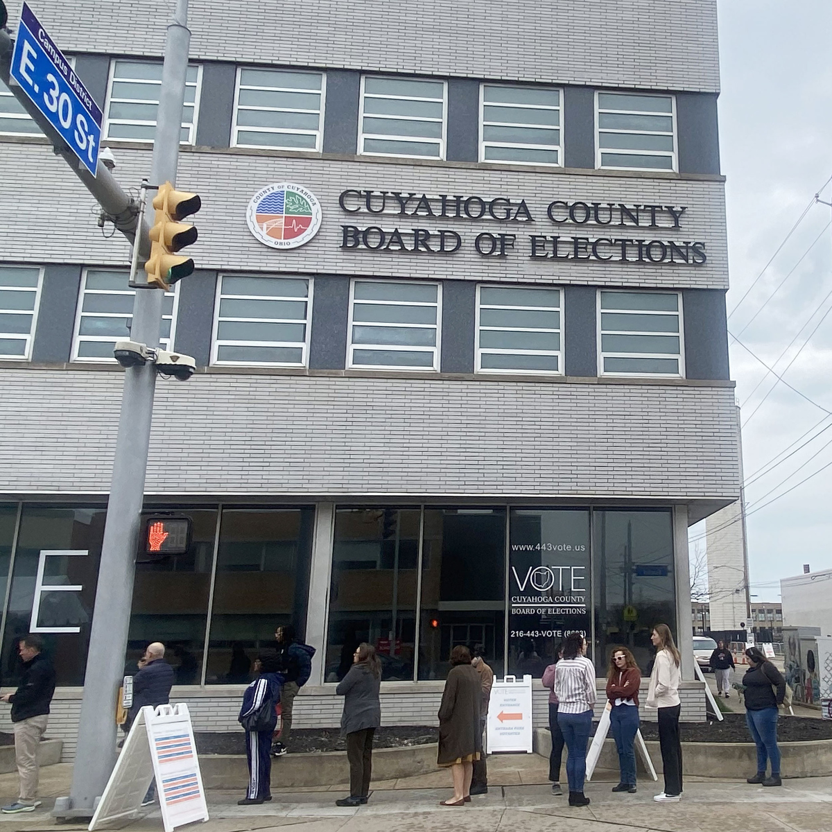 A photograph of more than 10 people lining up outside a gray building with a sign reading “Cuyahoga County Board of Elections.”