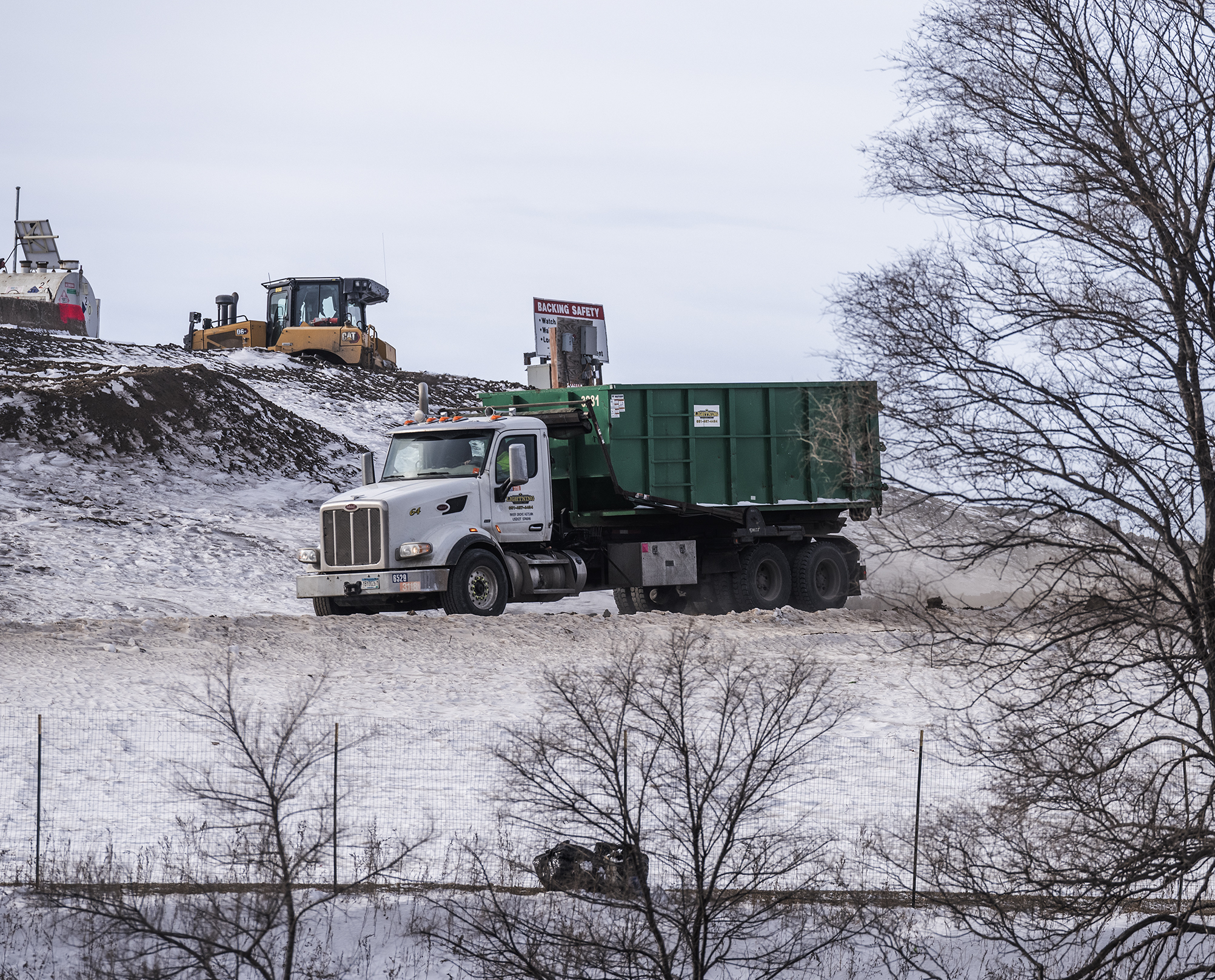 A white and green dump truck on a road with snow on the ground and other equipment in the background