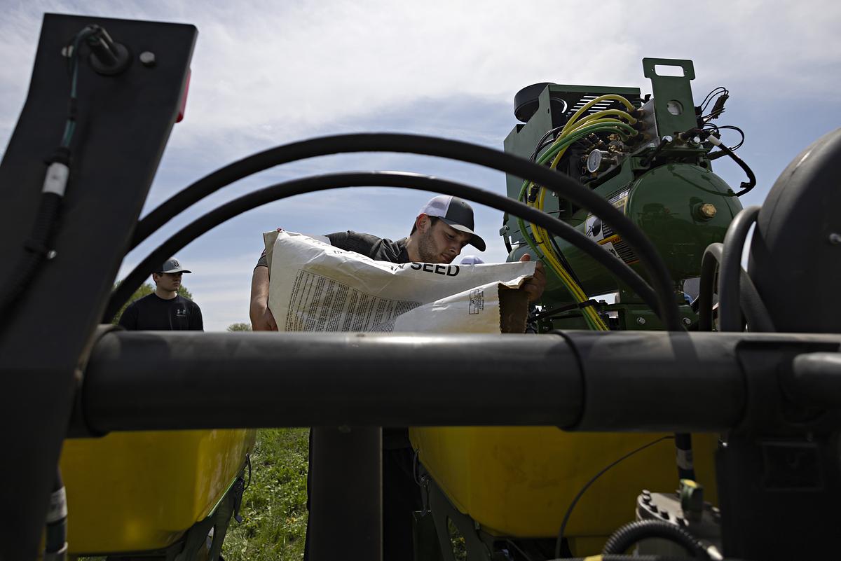 Young man in hat holds seed bag next to farm equipment.