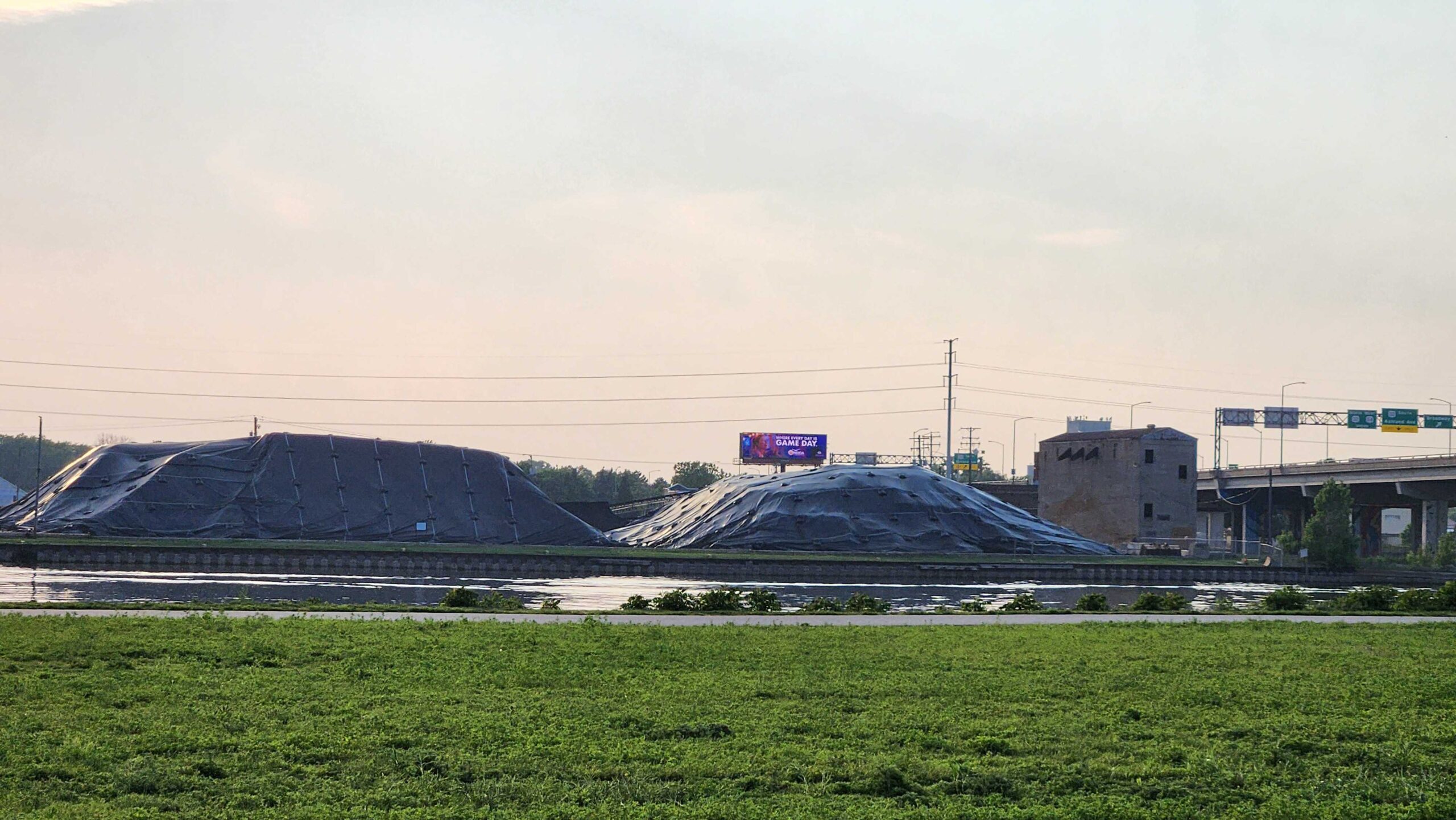 Tarped over mounds sit behind green grass and a river, bordered by a highway.