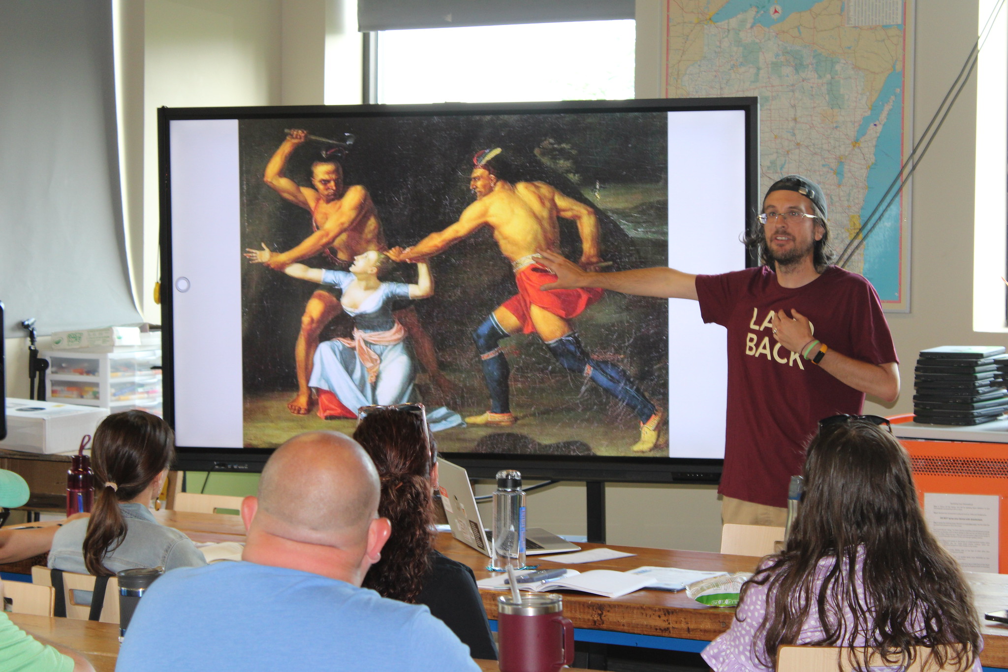 Seated people watch as a man stands facing them with his arm and hand extended over an image of Native Americans.