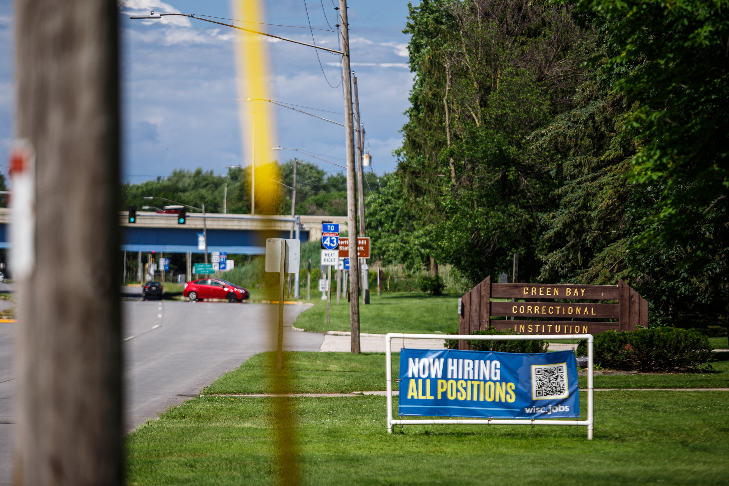 Sign says “NOW HIRING ALL POSITIONS” in front of sign that says “GREEN BAY CORRECTIONAL INSTITUTION” next to highway.