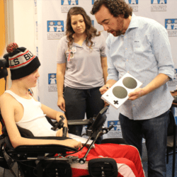 Mark Barlet showing a young man in a wheelchair an xbox adaptive controller while a young woman in a purple polo shirts looks on