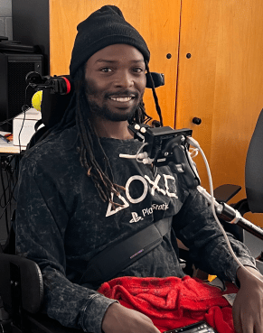 an image of a young man wearing a black stocking cap and dark grey long sleeve tshirt there is a QuadStick mounted in front of him