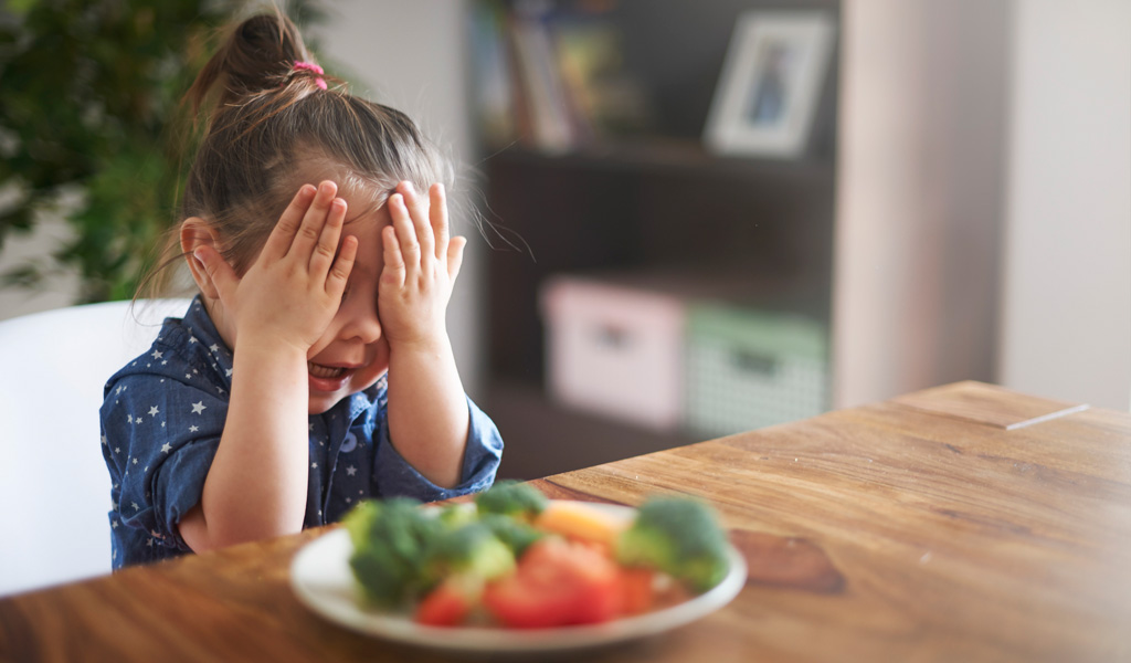 Girl with ADHD covers face with hand while sitting in front of plate of vegetables