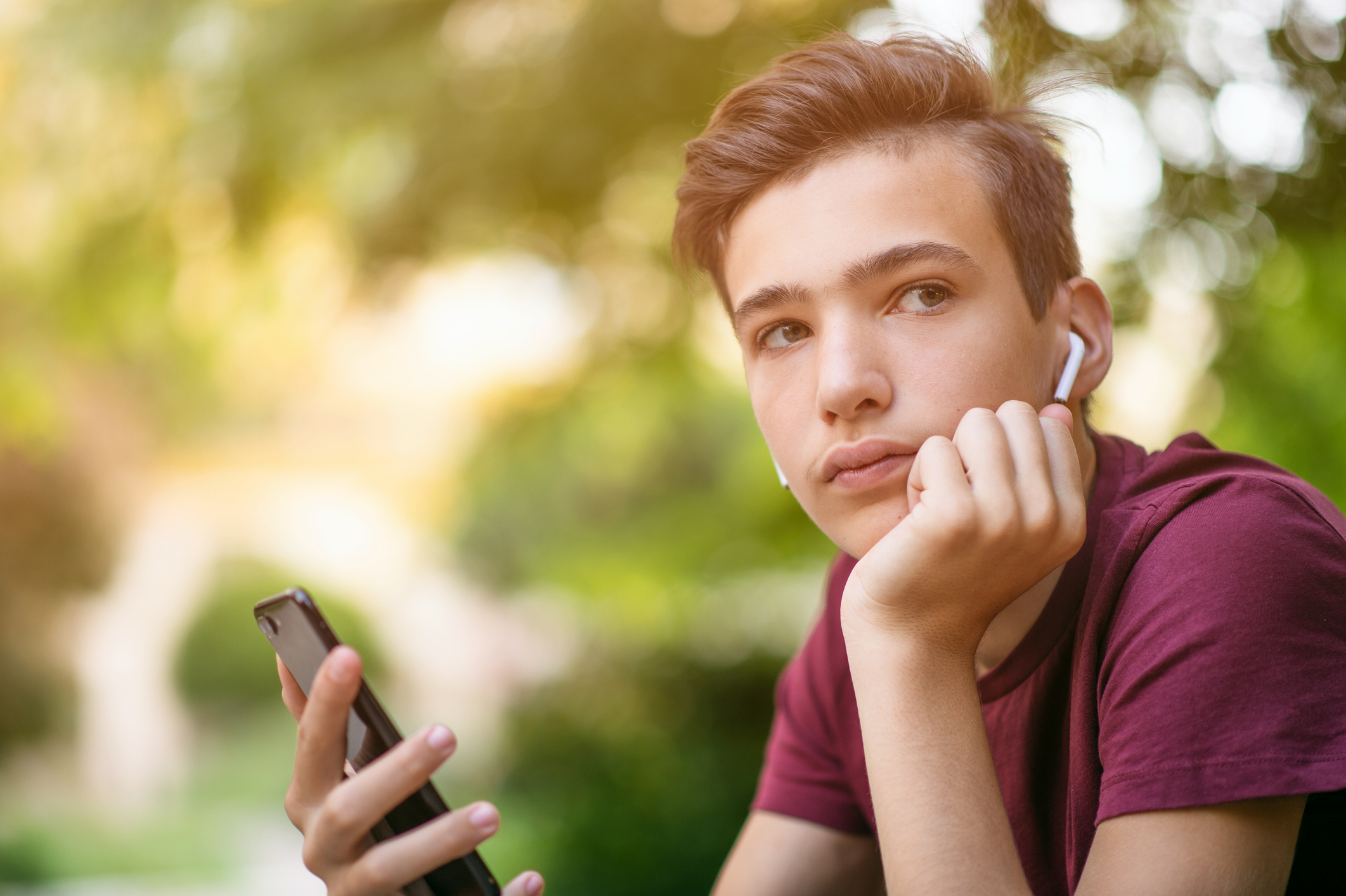 Close-up portrait of a thoughtful unhappy teenage boy with smartphone, outdoors.  Sad teenager with mobile phone looks away, in the park.  Pensive teenager in casual clothes with cell phone in park