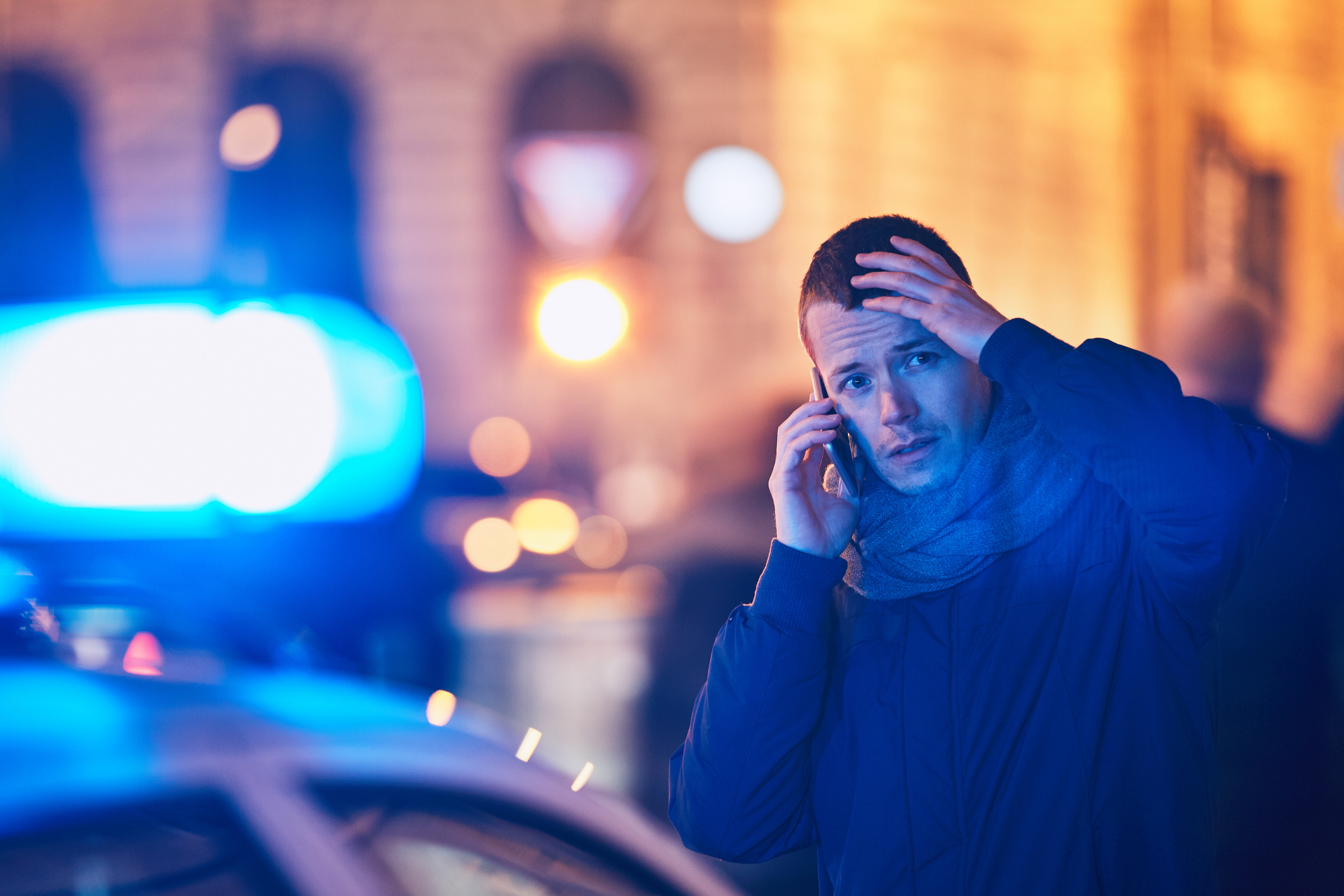 A young man is on the phone after a crisis situation on a city street. Police lights can be seen in the background.