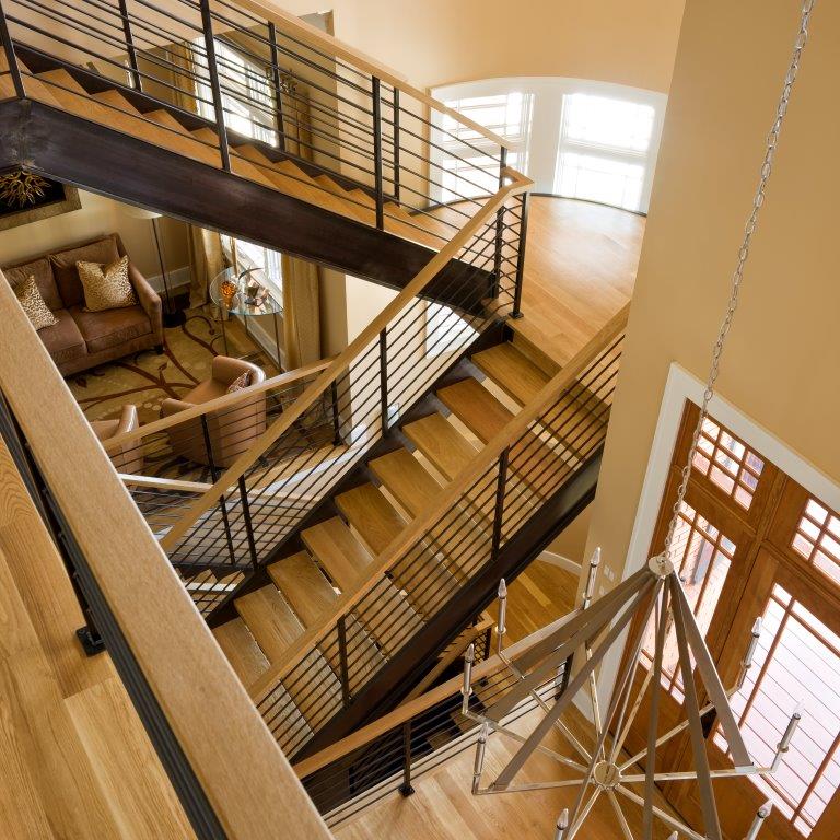 The chic staircase is the centerpiece of this house, even visible from the outside intentionally through the placement of the windows. The railing in a combination of warm wood and metal cable is an excellent example of Industrial Modern Farmhouse. The light fixtures were chosen because they feature both brushed nickel and wood. The color scheme is neutral beige with white trim to enhance the openness of the foyer and sitting area. (Sharon McCormick Designs, Gordon Beall Photography)
