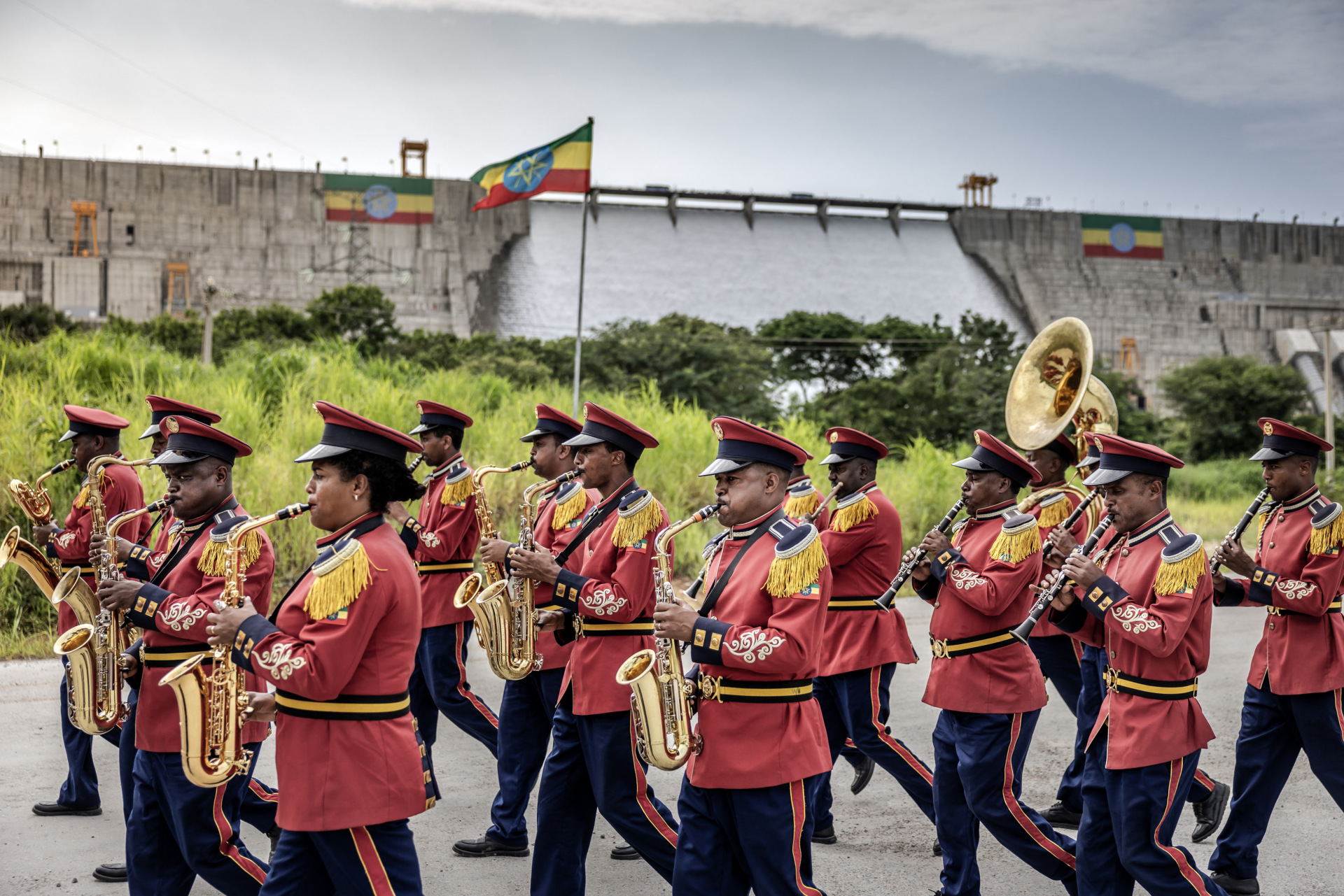 The band members march toward the left of the photo, wearing red jackets and black pants with red stripes and playing instruments including a tuba, as a red, yellow and green Ethiopian flag flies in the background in front of the dam, with water flowing down its slope.