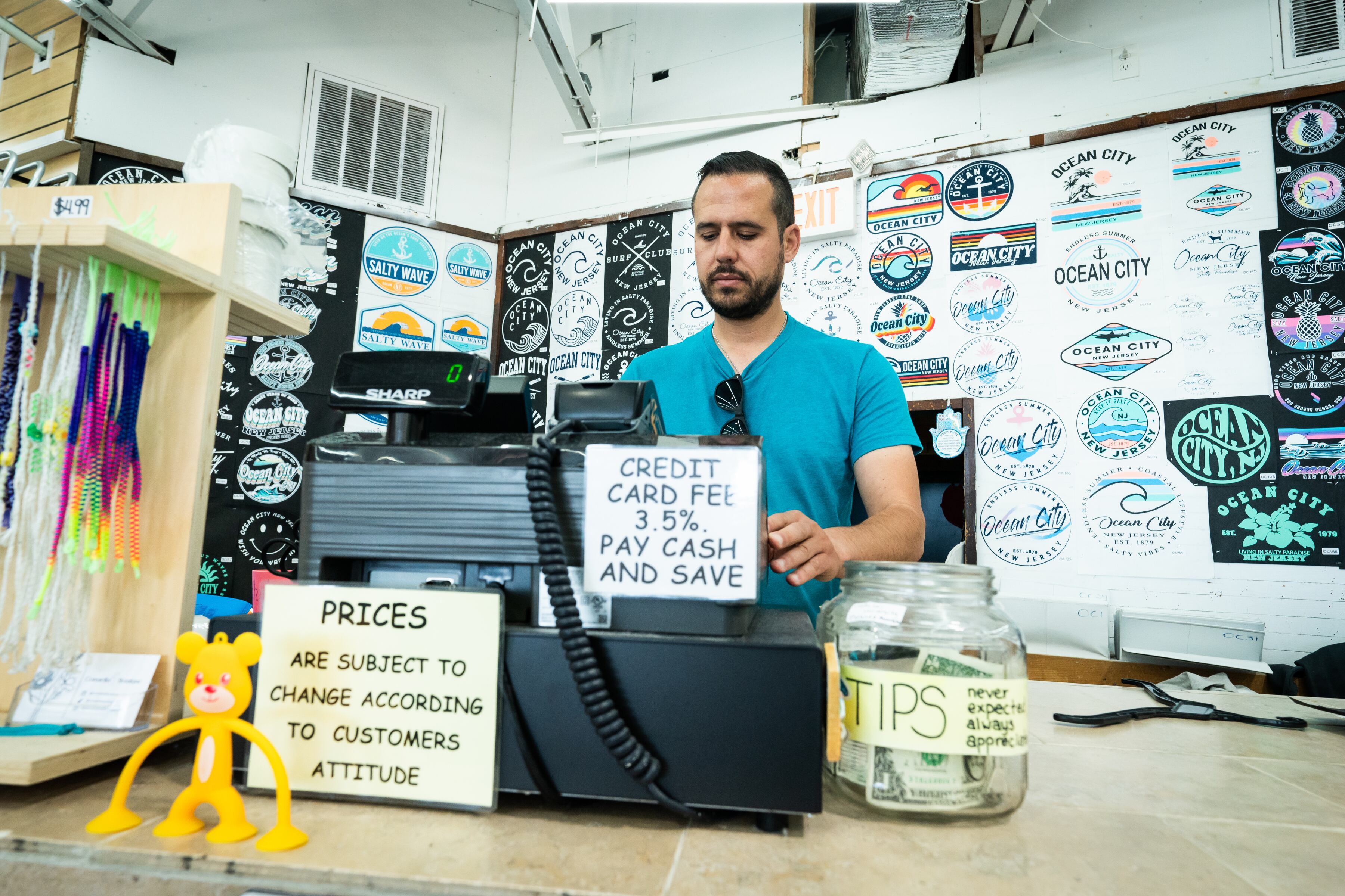 Eli Romy mans the register at one of his boardwalk shops, Shelly's by the Sea, in Ocean City. A sign tells customers they will pay more if they use a credit card. (Jessica Griffin / The Philadelphia Inquirer)