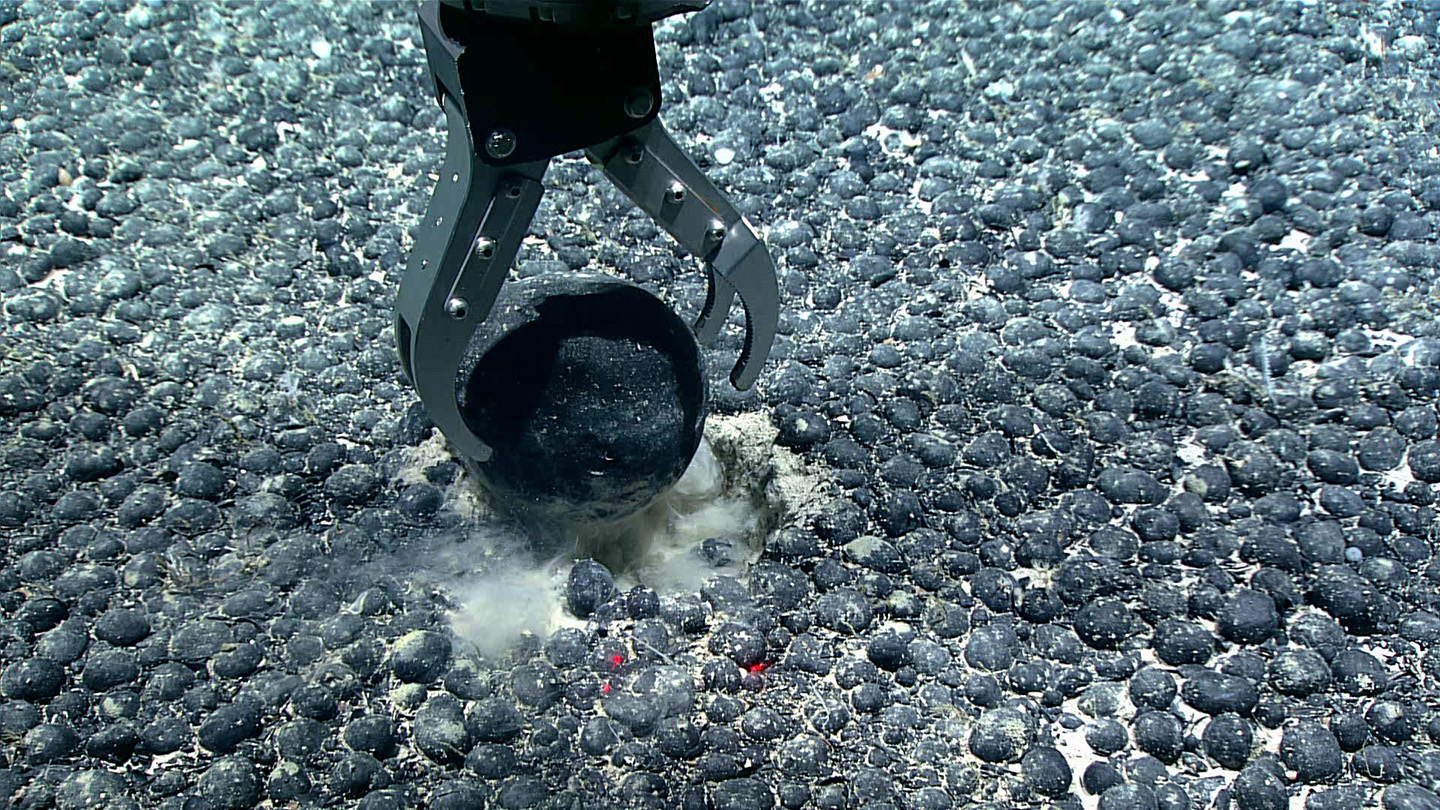A metal claw reaches for a round nodule on the seafloor.