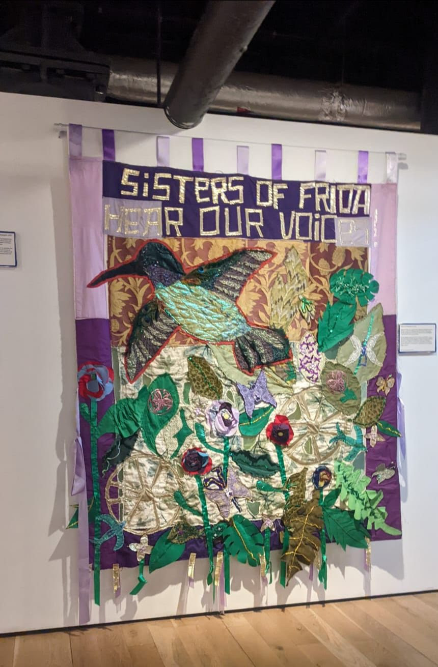 SOF banner hanging from wooden pole. Multi colours back ground light purple and dark purple. Visible a bird, leaves, flowers, 2 wheels, feathers. Text on banner - 'Sisters of Frida Hear Our Voices'