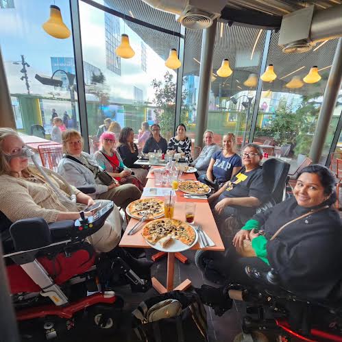 group of women around a long table, with 3 wheelchair users. one has a breathing tube, some have pizzas in front of them.