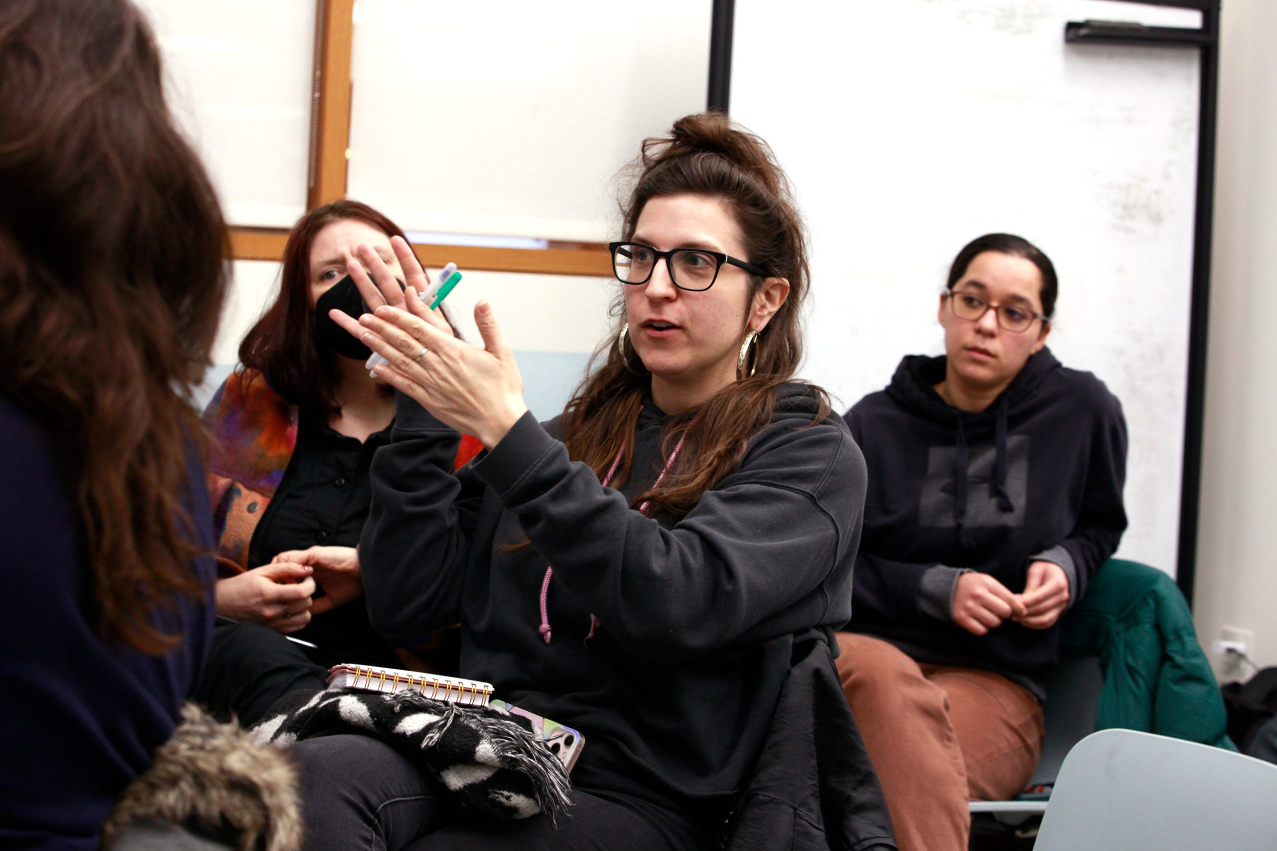 Three women sit inside a library room as one speaks and gestures with her hands.