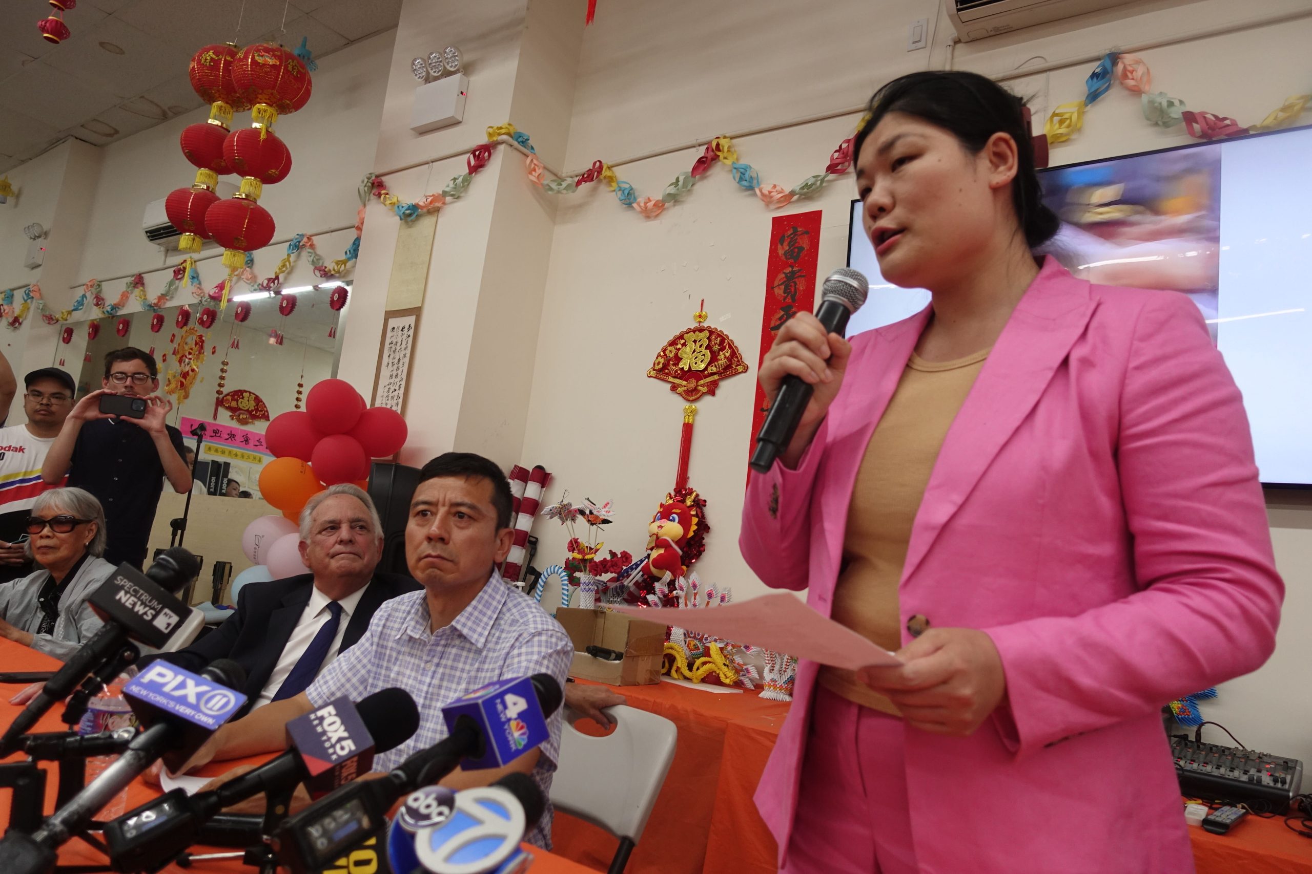 Councilwoman Susan Zhuang stands holding a microphone in a pink suit and tan T-shirt. The left side of the image shows network news microphones and community attendees.