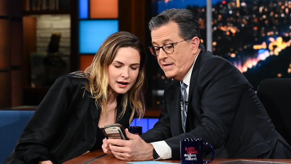 A woman with light-toned skinned and a man with light-toned skin, each in blazers, look at a phone held by the man. The people are Rebecca Ferguson and Stephen Colbert on the "Late Show" set.