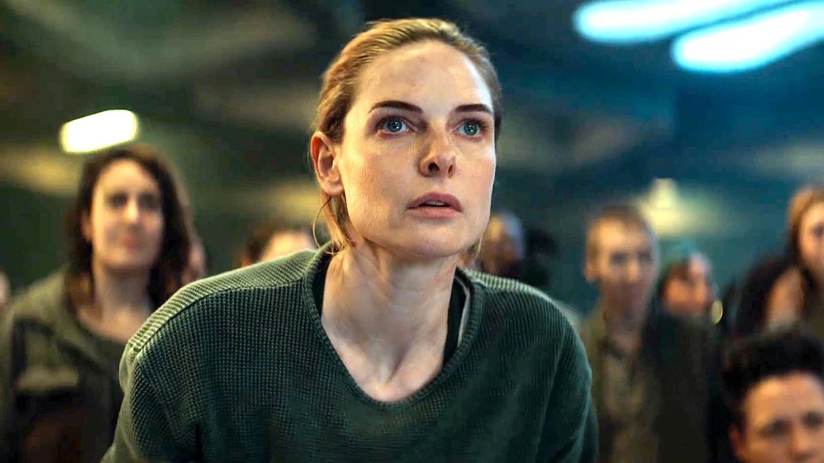 Rebecca Furguson stars in Silo. A woman with brown hair pulled back, wearing a basic green sweater stares out of frame