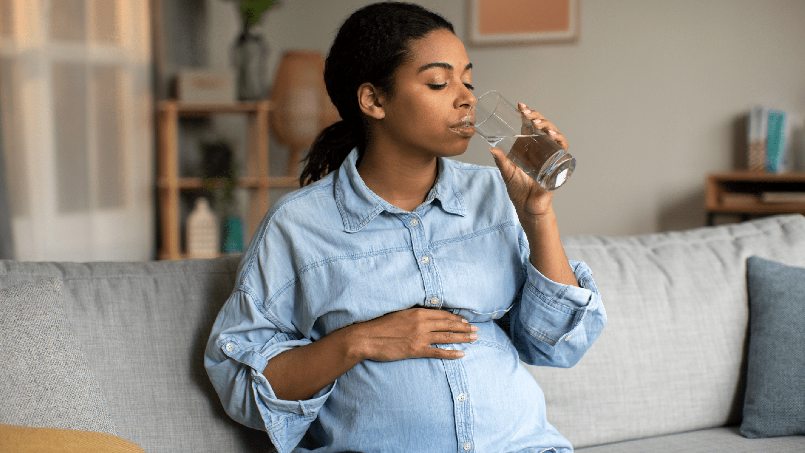 A pregnant, African-American woman in a blue shirt drinks water on a grey couch.