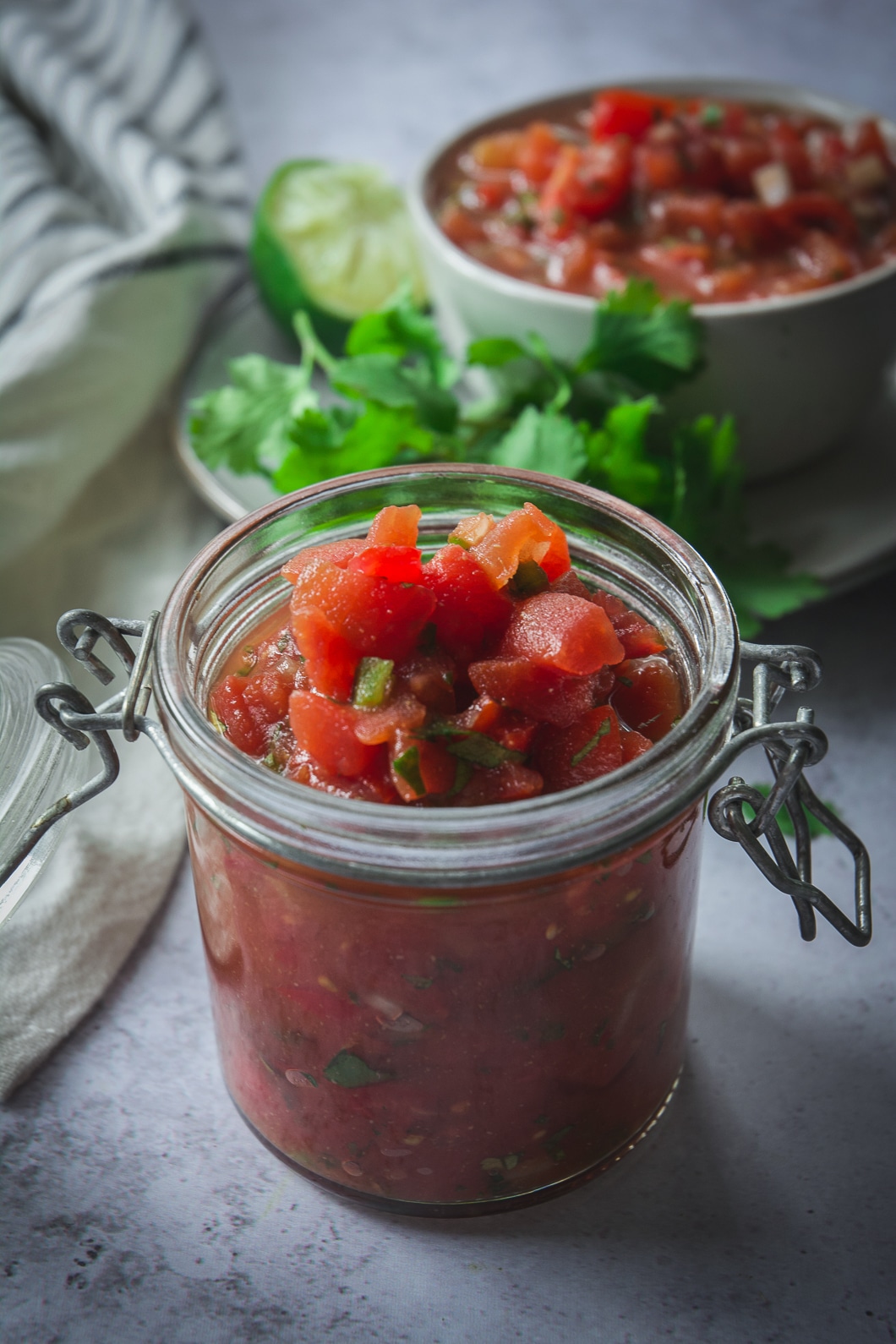 Homemade salsa in jar with cilantro and bowl of salsa in the background