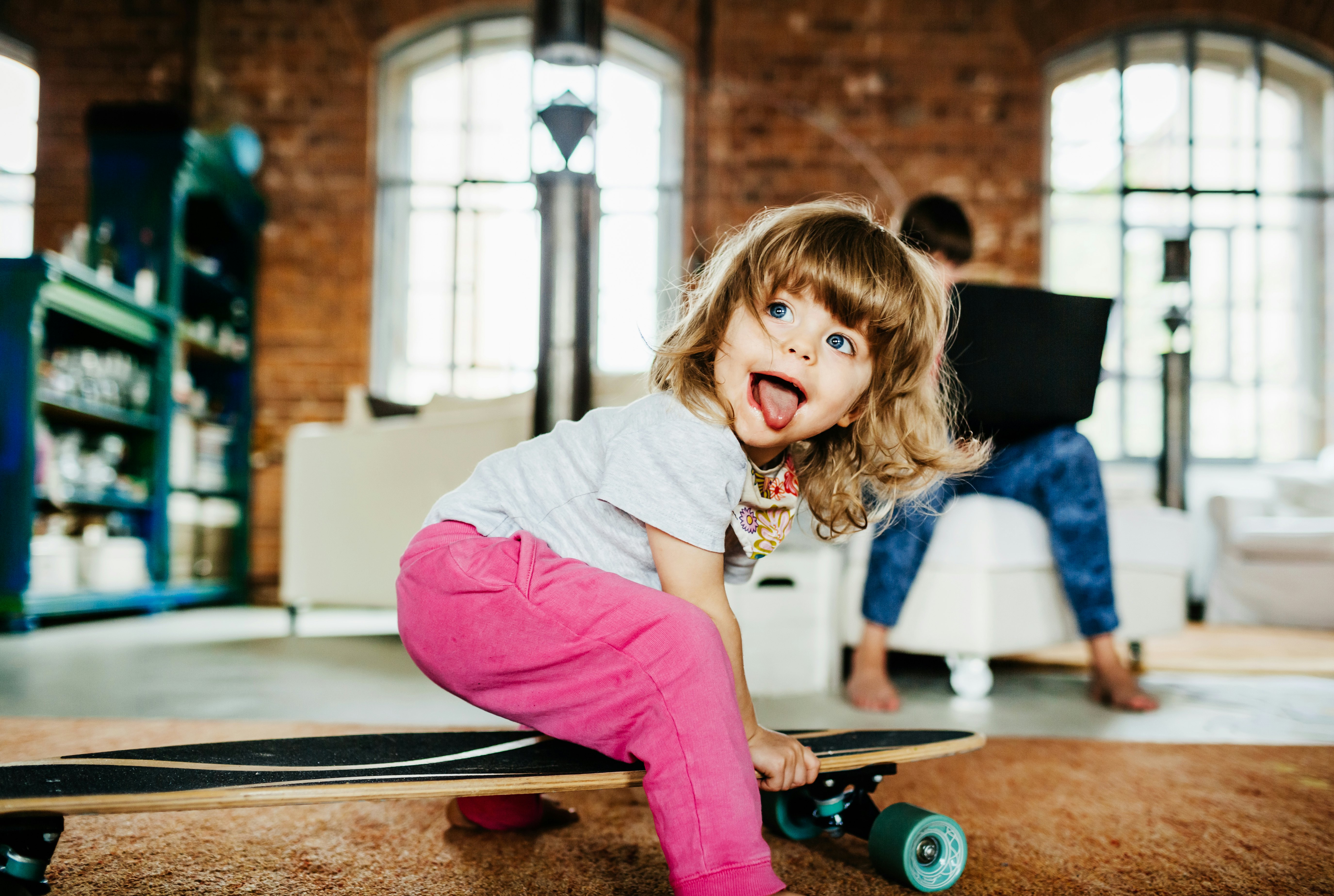 A toddler playing on a skateboard indoors while her mother works on a laptop.