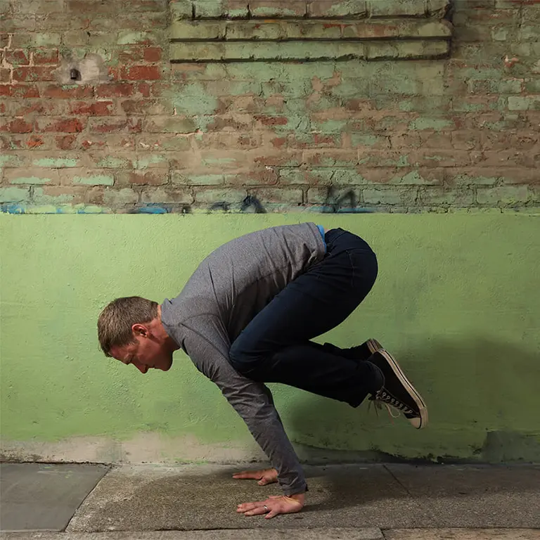 Jason Crandell in Crow pose against a green brick wall
