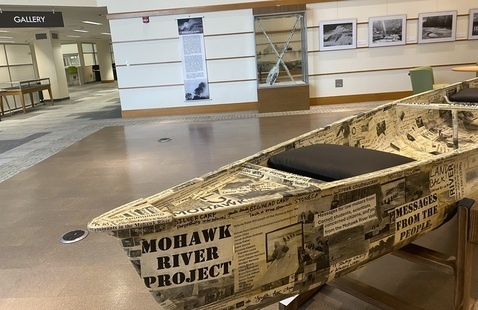 Photo of a canoe decorated with messages in the foreground and a background of photographs and two paddles in glass case. 