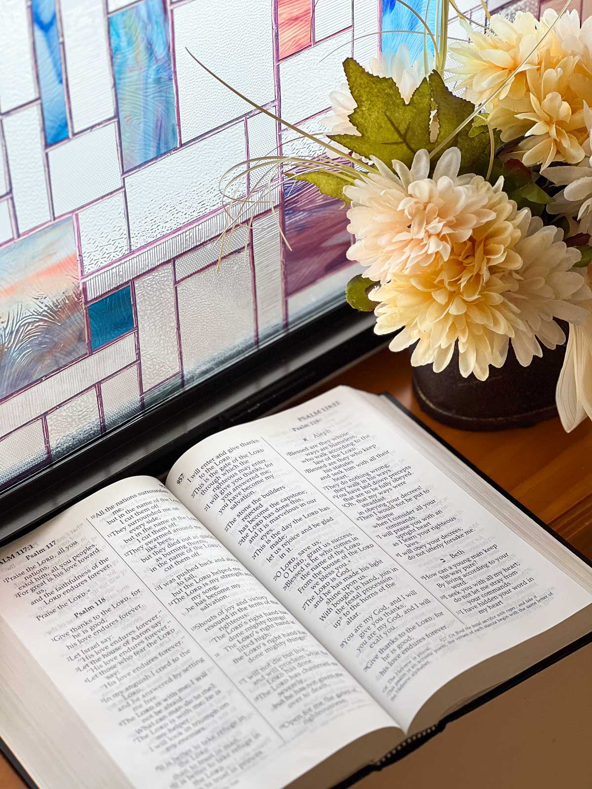 open Bible on a windowsill with a stained glass window and flower arrangement.