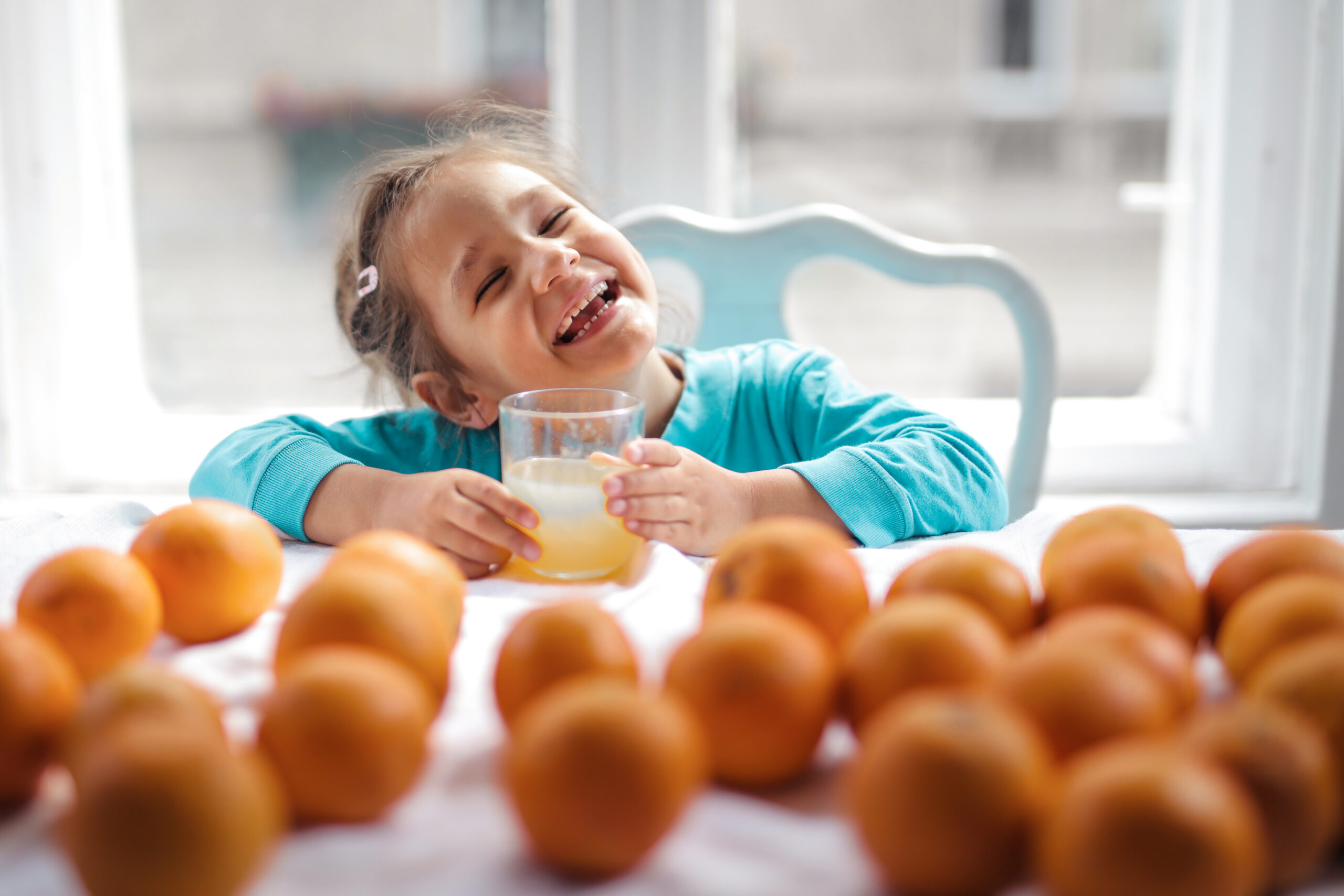 girl sitting at table with glass of orange juice and smiling. Oranges in foreground on table as illustrating healthy foods to eat during a mental health day