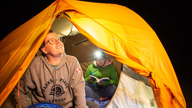 A young man looks out of his tent while a women reads a book at Oster Lake Backcountry Campground in Elk Island National Park.