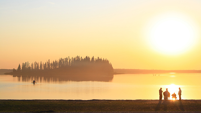 Silhouettes of a group of people on Astotin Beach at sunset. An island and a paddler are visible in the background. 