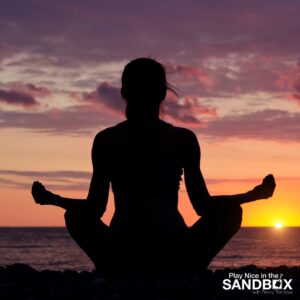 image of a woman from behind sitting on a beach at sunrise in a cross legged yoga pose