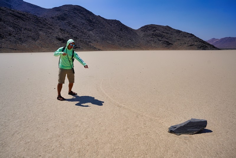 David Stock Jr of Divergent Travelers Adventure Travel Blog standing a pointing at a racetrack rock at Racetrack Playa in Death Valley National Park.
