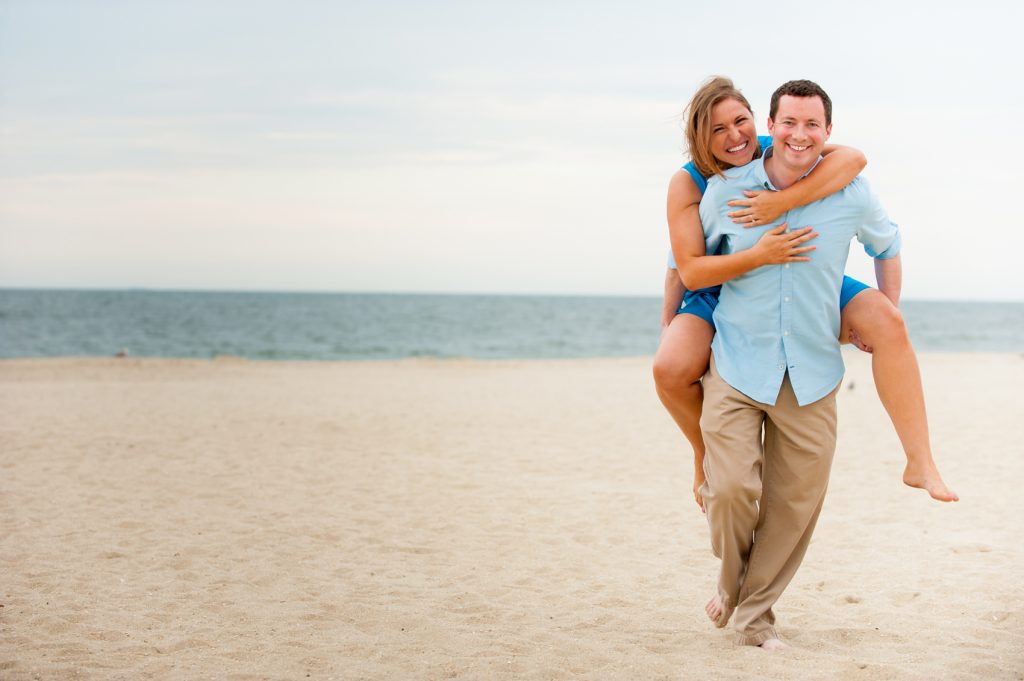 couple piggybacks on the shoreline of in New Jersey during their beach engagement photoshoot