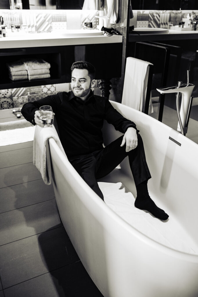 groom sits on the bath tub while holding a glass of champagne