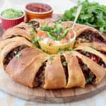 Taco ring with crescent rolls on wood cutting board with ramekins of sour cream, salsa and guacamole