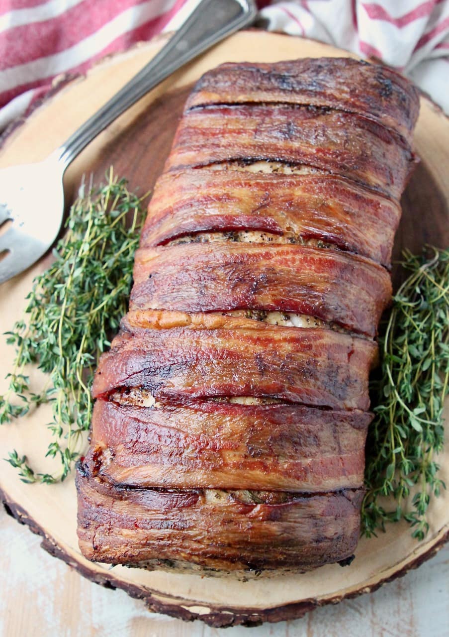 Bacon wrapped pork loin with herbs on wood cutting board with large serving fork