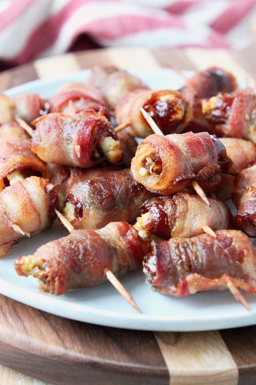Bacon wrapped dates piled up on white plate on top of wood cutting board