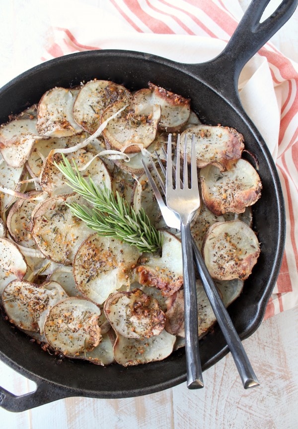 A cast iron skillet on a wooden surface filled with sliced and seasoned potatoes and two forks.